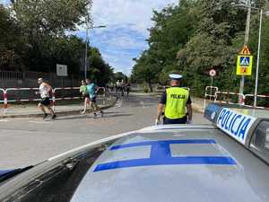Policjant stojący przy radiowozie patrzy na biegnących zawodników.