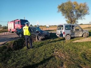 Zdjęcie przedstawia dwa rozbite samochodu przy skrzyżowaniu. Za nimi stoi radiowóz policyjny, a obok wóz bojowy straży. Przed rozbitymi samochodami stoi policjant w odblaskowej kamizelce