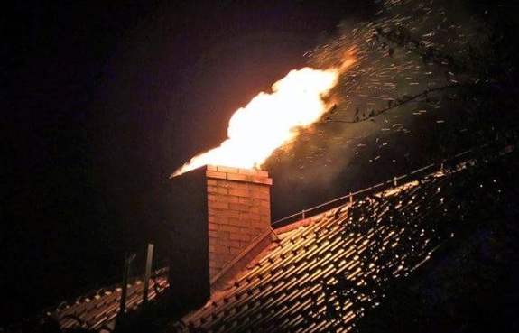 Grafika przedstawia fragment dachu z kominem. Z komina wydobywa się ogień i iskry