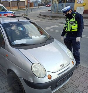 Umundurowany policjant ruchu drogowego w białej czapce i odblaskowej kamizelce przygląda się srebrnemu pojazdowi marki Daewoo Matiz