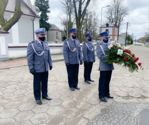 Czterech policjantów w umundurowaniu galowym stoi przed pomnikiem. Jeden z policjantów trzyma biało - czerwoną wiązankę