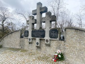 Zdjęcie przedstawia pomnik złożony z trzech krzyży. Po prawej stronie leży na ziemi wiązanka i stoją dwa znicze