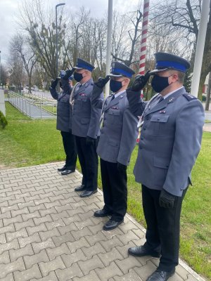 Czterech policjantów w umundurowaniu galowym salutuje przed tablicą umieszczoną na budynku sochaczewskiej komendy