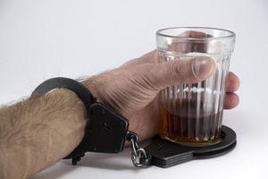 Męska w ręku trzyma szklankę z drinkiem. Kajdanki założone są na rękę i szklankę.
