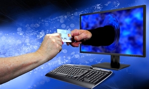 Dwie dłonie, jedna wyciągnięta z monitora, obie trzymają kartę bankomatową.