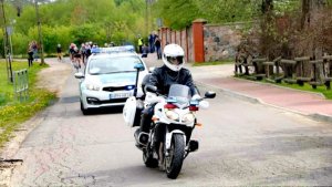 Wizualizacja przedstawia policjanta ruchu drogowego na motocyklu, policyjny radiowóz oraz grupę kolarzy.