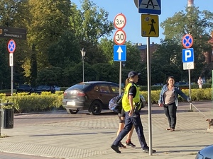 Obraz przedstawia ulice Żyrardowa, na zdjęciu widoczna jest policjantka przeprowadzająca dziecko przez przejście dla pieszych